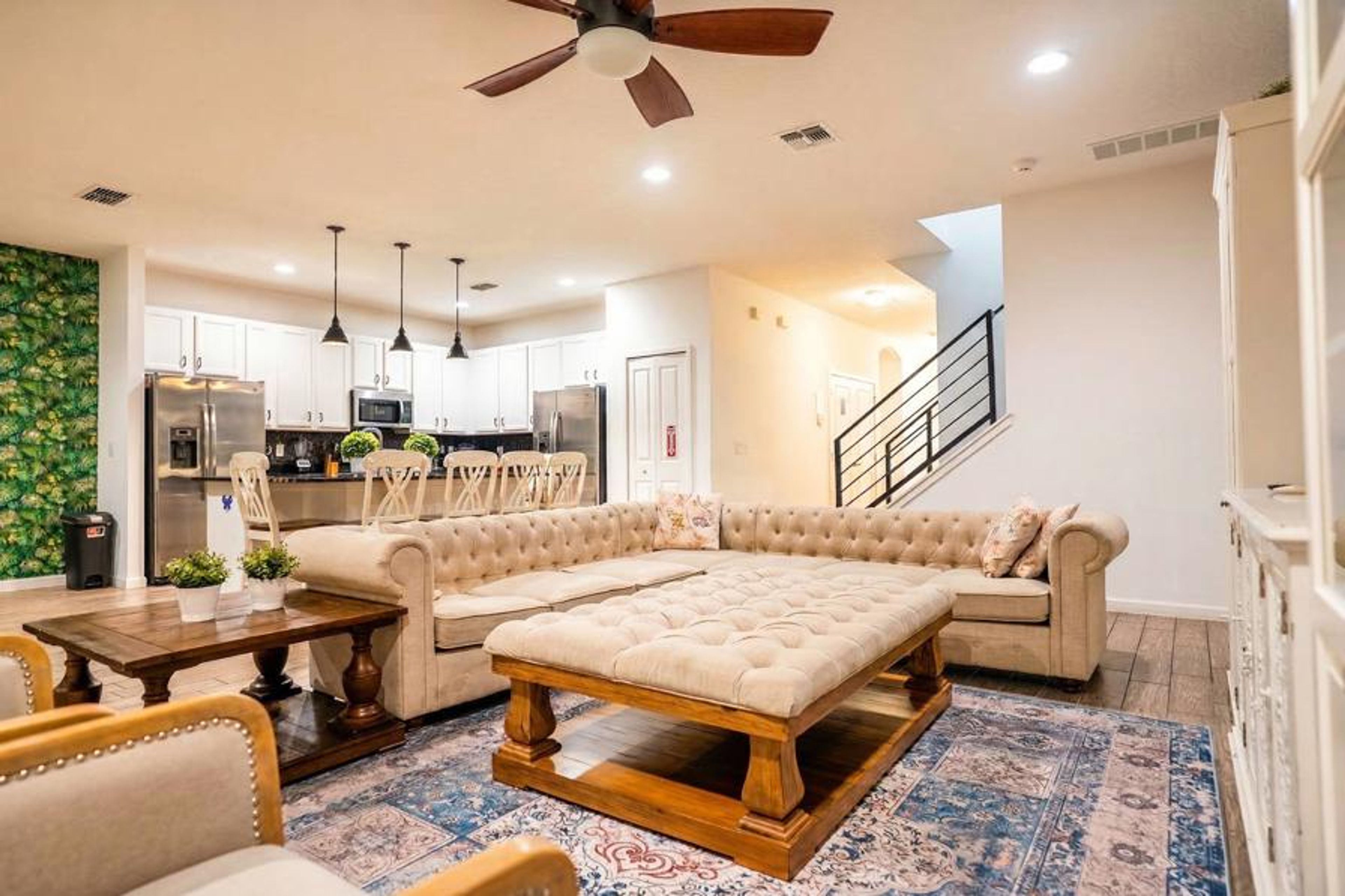 Relax in an elegant open living area with a plush sectional sofa, tufted ottoman, wood accents, and a bright kitchen in the background. Warm lighting, soft tones, and stylish details create a comfortable space for family time.