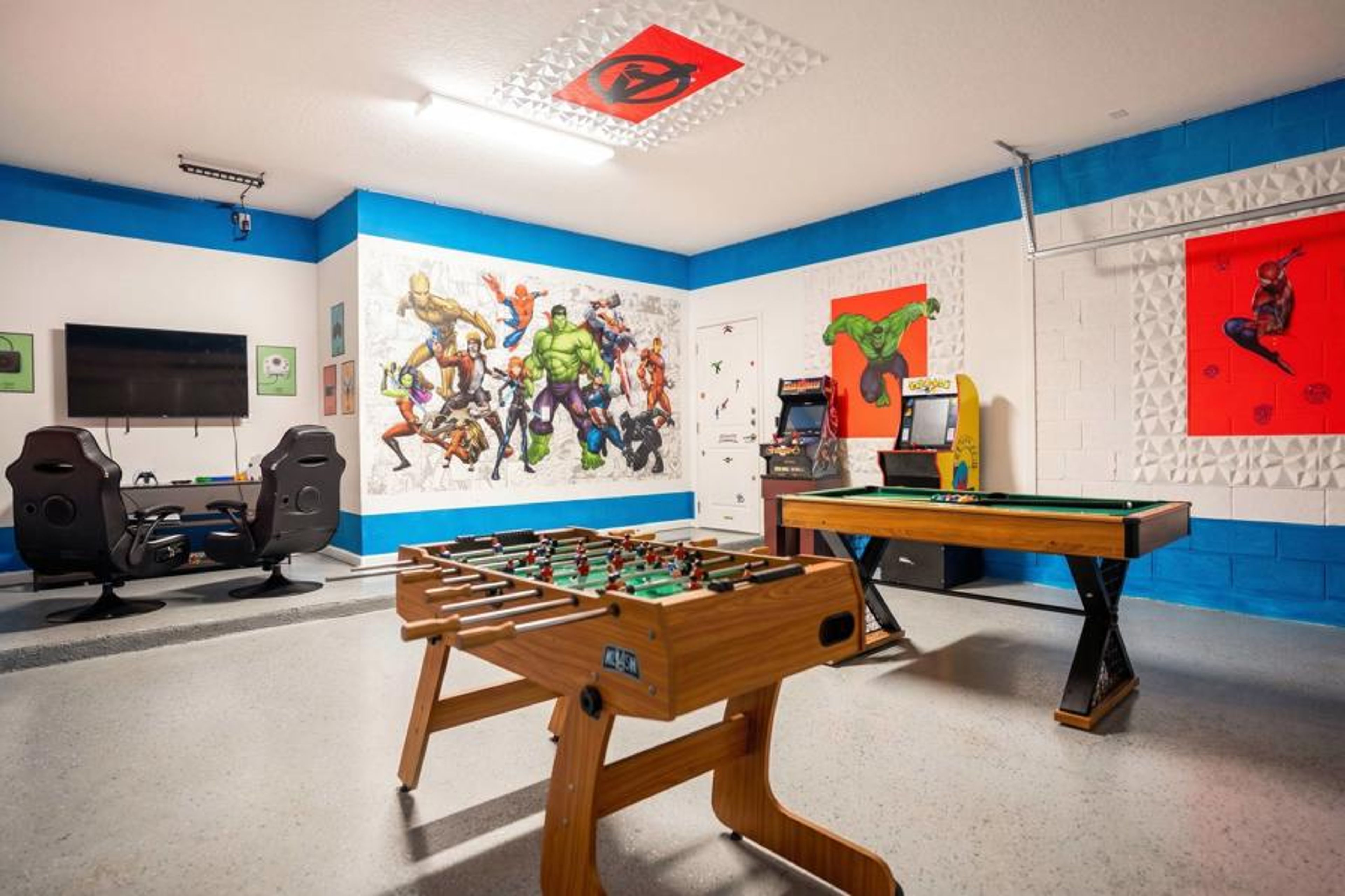 Enjoy a bold superhero-themed game room with foosball, pool table, arcade games, gaming chairs, and a wall-mounted TV. Colorful murals and bright blue accents create a fun space for kids, teens, and adults to play together.