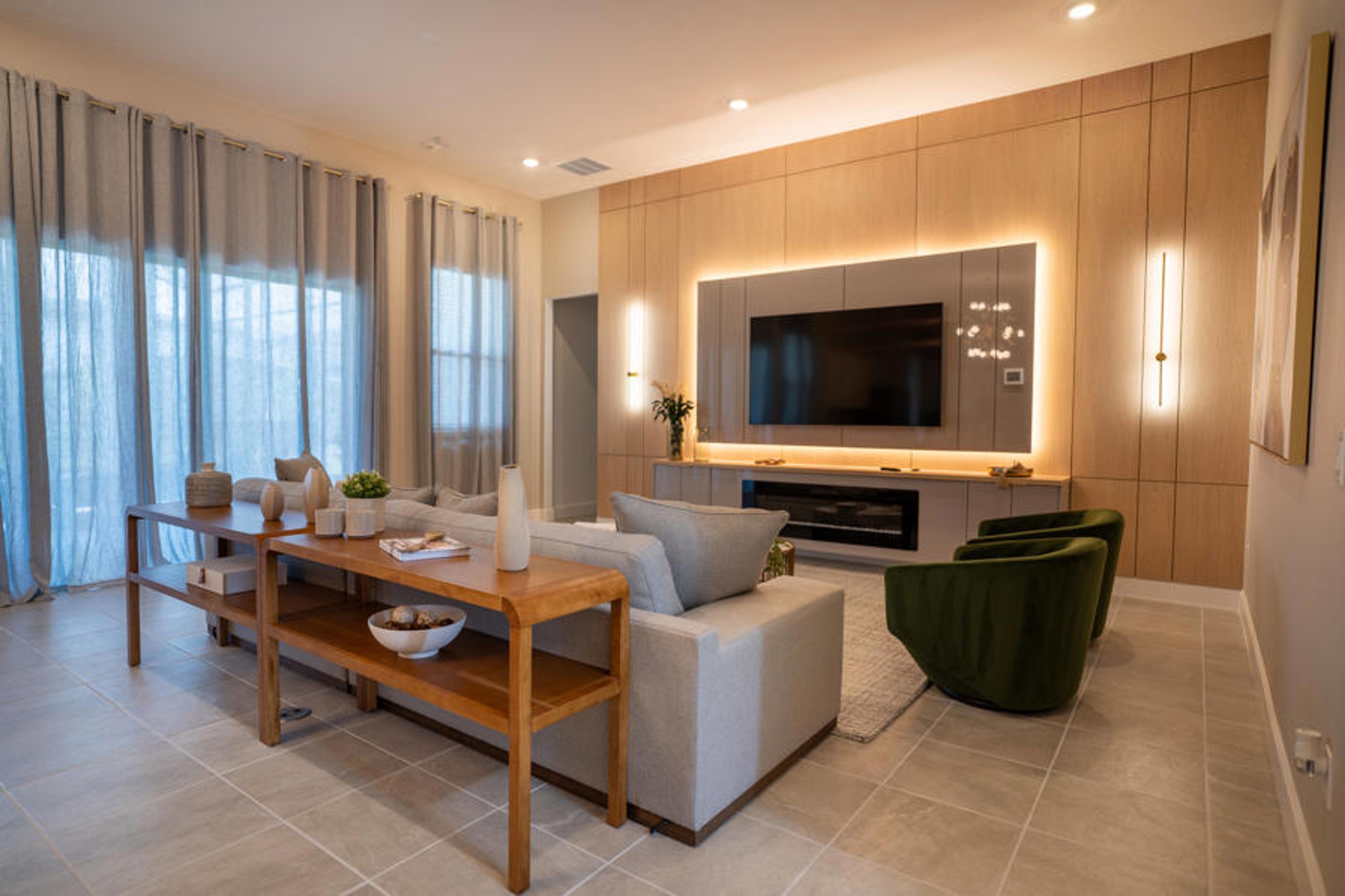 Experience a stylish living area with a modern entertainment unit and warm wood accents. The soft neutral furniture and gentle wall lighting create a relaxing atmosphere, while large windows brighten the space with natural light.