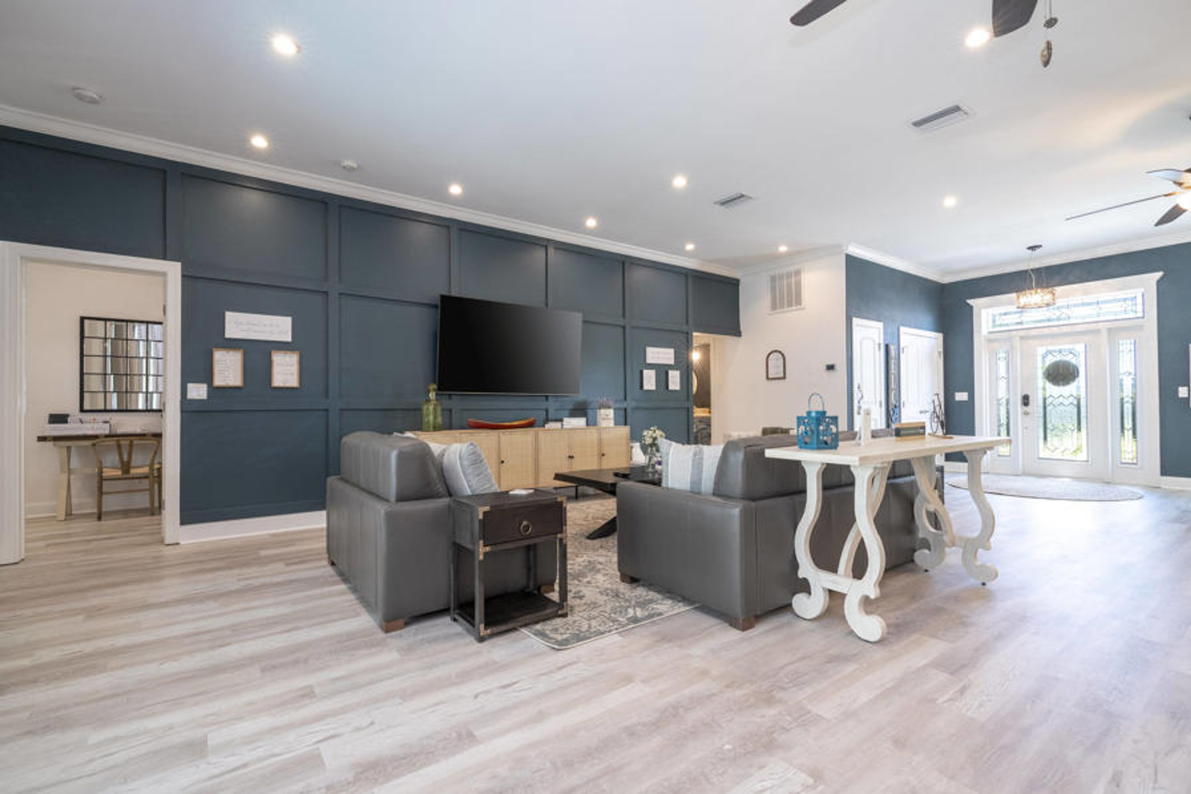 Enjoy the spacious living area with a striking dark blue accent wall that contrasts beautifully with light wood flooring. Relax on stylish gray sofas, and soak up the abundant natural light streaming through large glass doors.