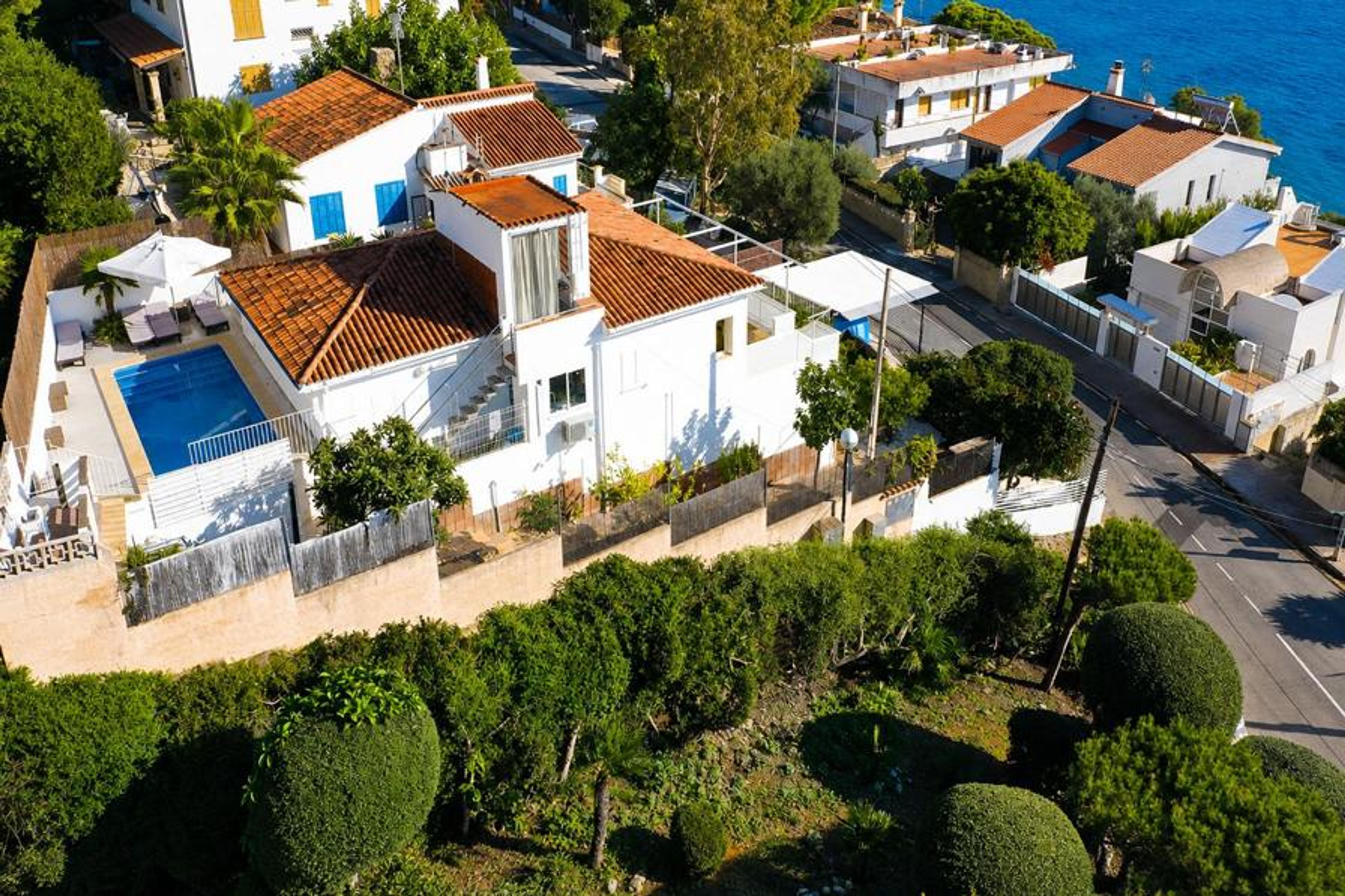 An aerial view showcases a multi-level white villa with traditional terracotta tile roofs, featuring a private swimming pool, sun loungers, and an elevated terrace just a short distance from the blue sea.