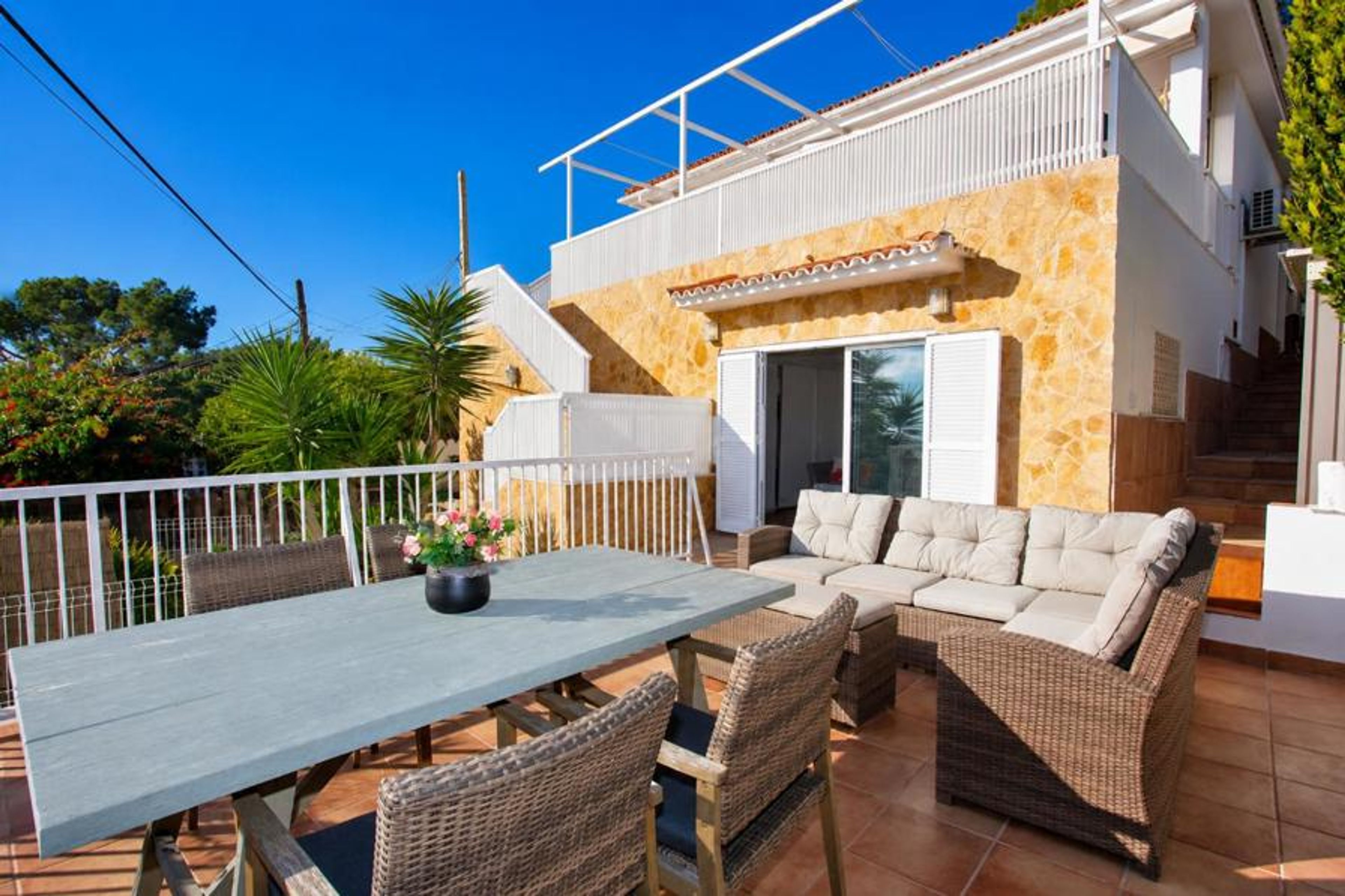 A spacious outdoor terrace features a wicker sectional sofa and a large dining table set against a stone-facade villa under a clear sky.