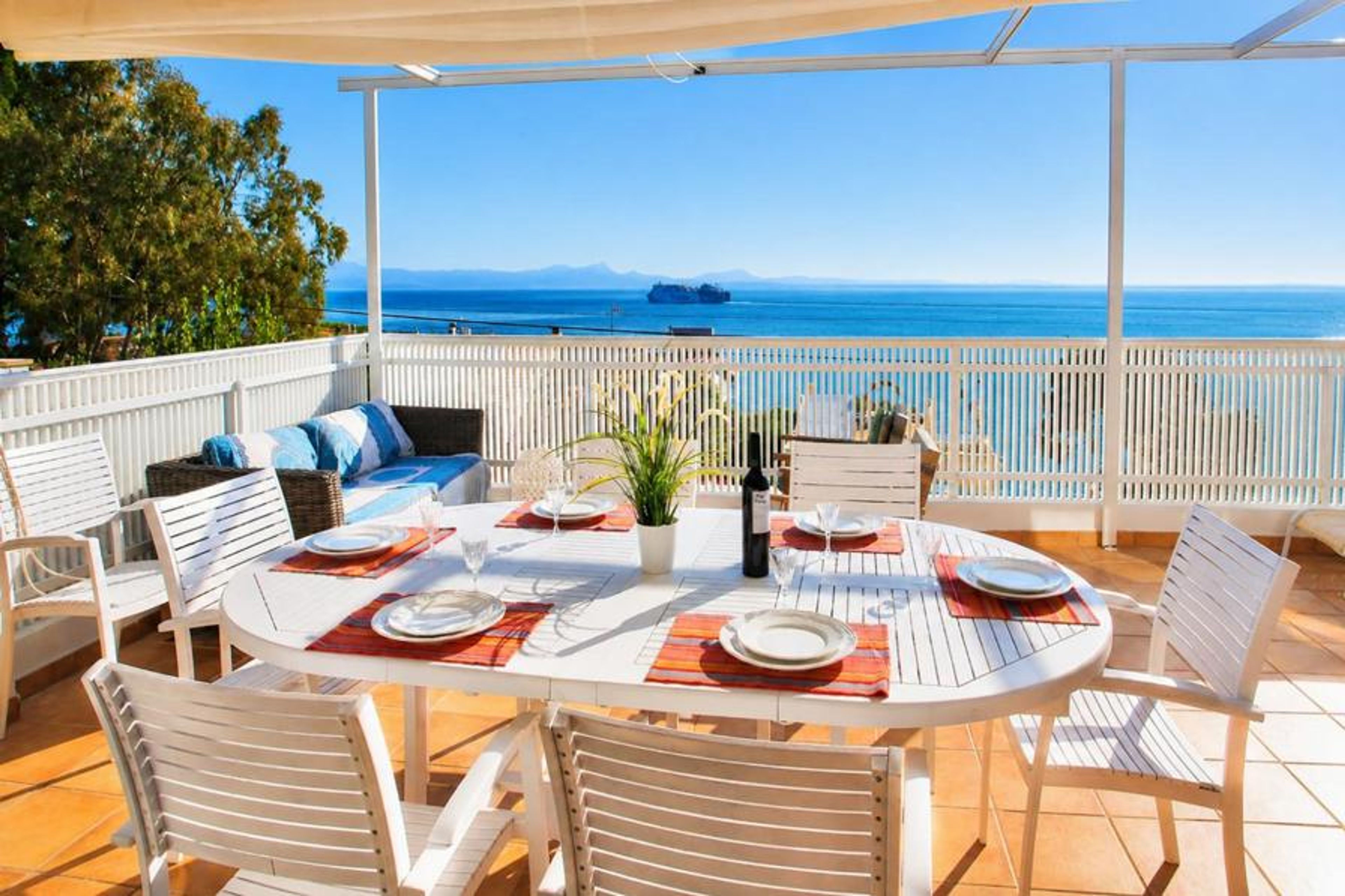 An outdoor dining table is set for a meal on a shaded rooftop terrace, offering sweeping panoramic views of the sea and distant mountains.