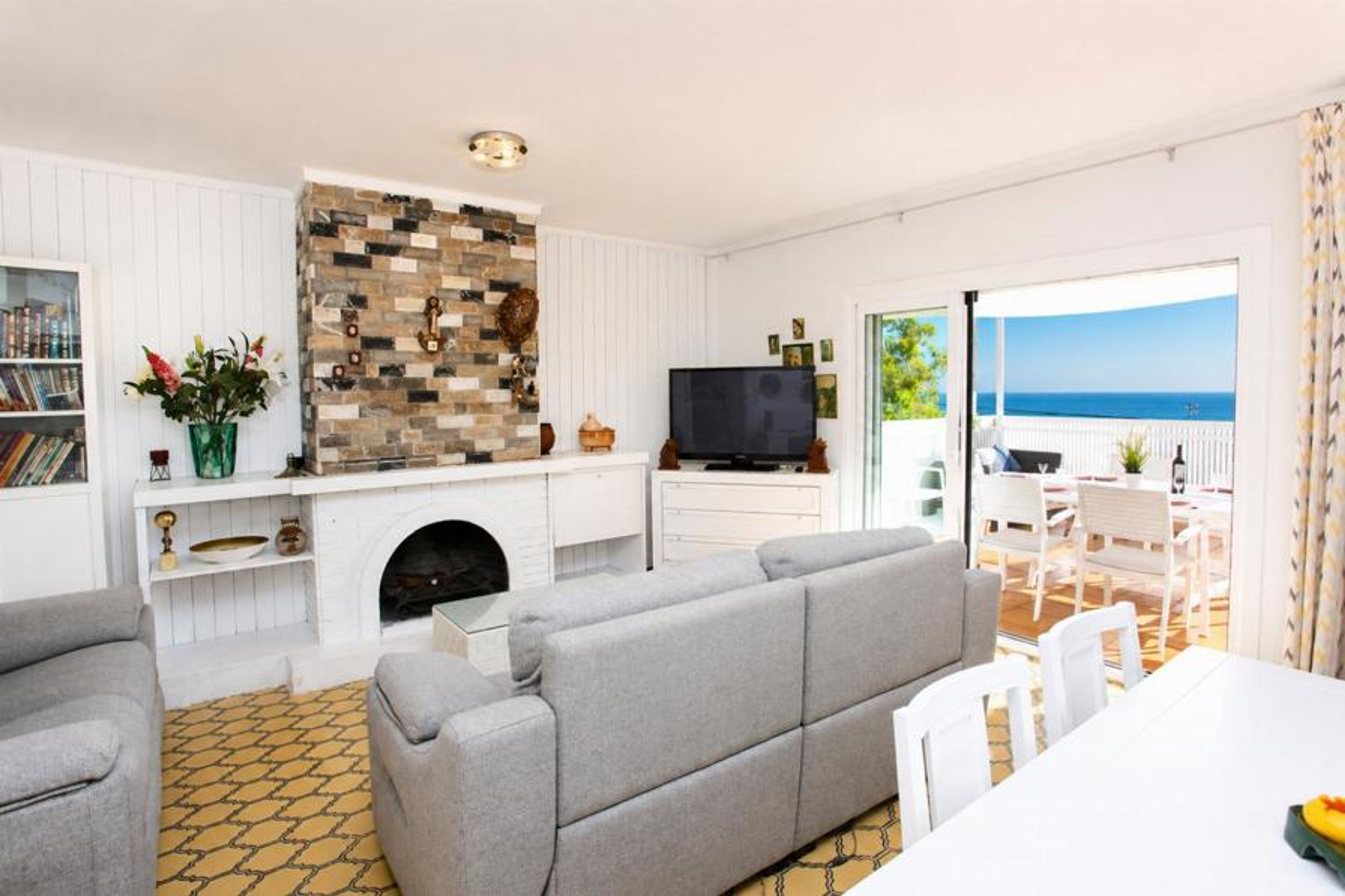 A bright living room features cozy gray sofas and a brick fireplace accent, with large sliding glass doors opening directly onto a sea-view dining terrace.