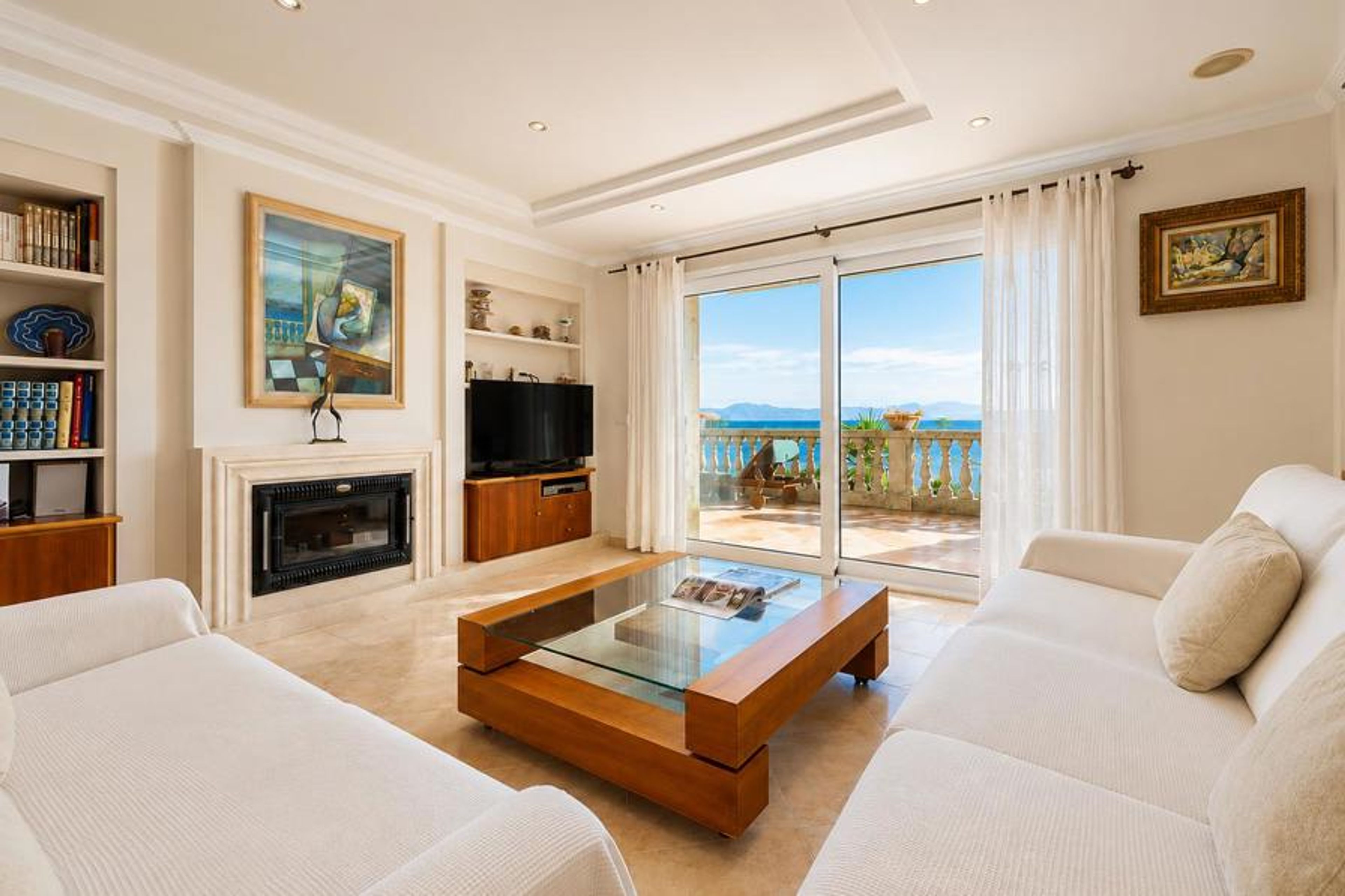 A bright, modern living space featuring cream sofas, a central fireplace, and large sliding glass doors that open to a scenic seaside terrace.