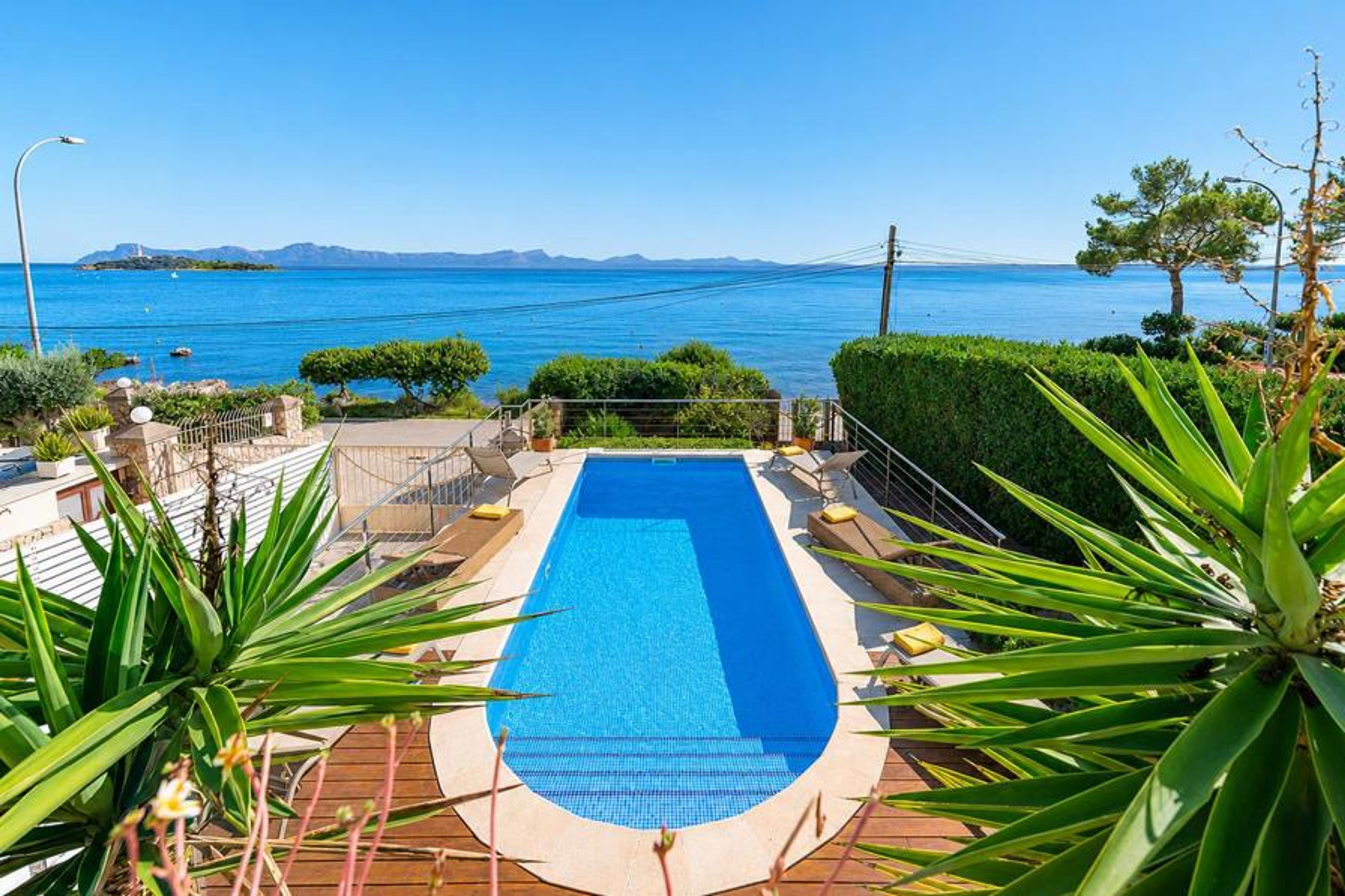A pristine, oval-ended swimming pool surrounded by a wooden deck, lounge chairs, and vibrant tropical greenery overlooking a calm ocean bay.