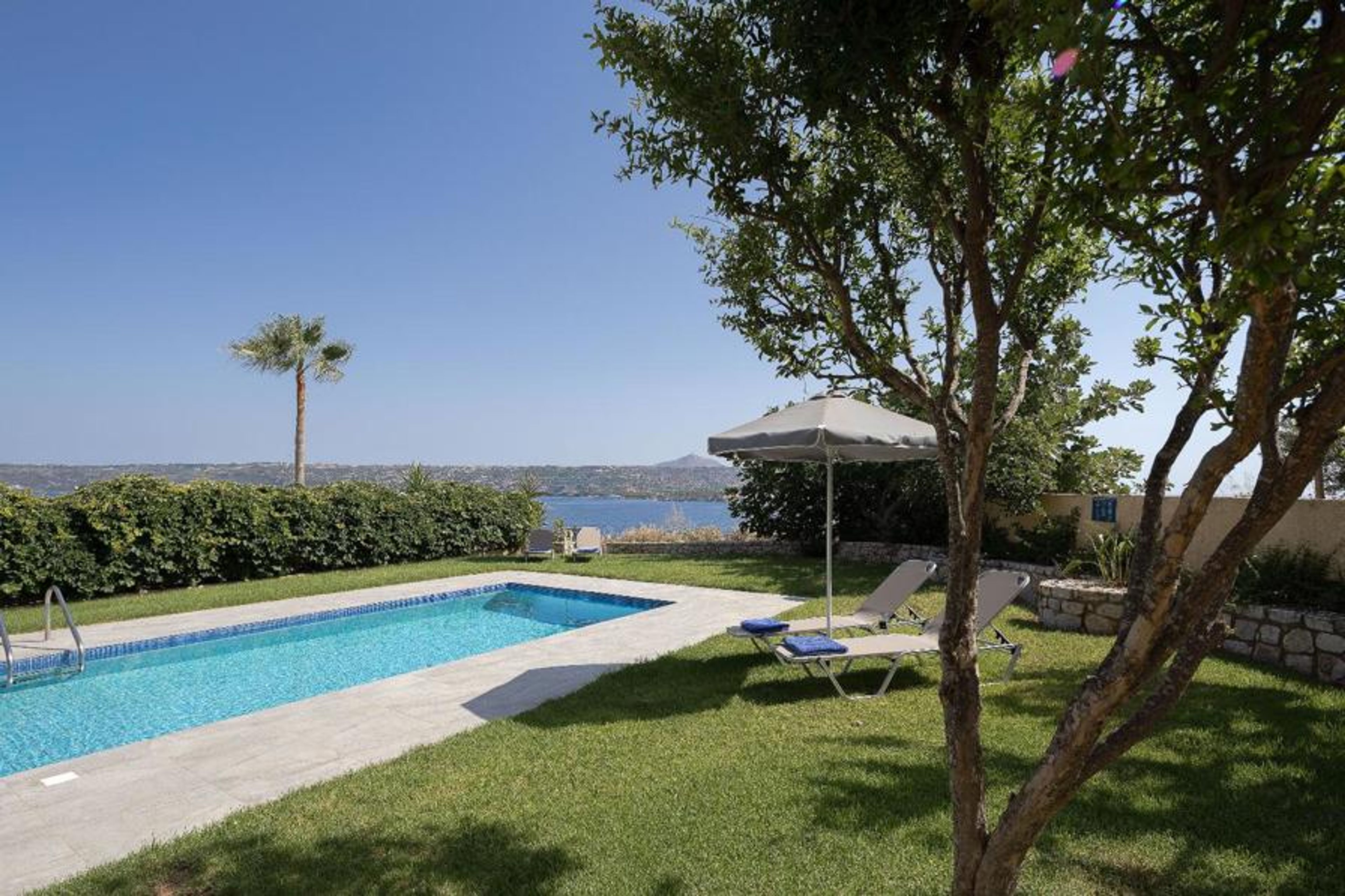 Private swimming pool with sun loungers, shaded garden seating, lush Mediterranean greenery, and beautiful sea views.