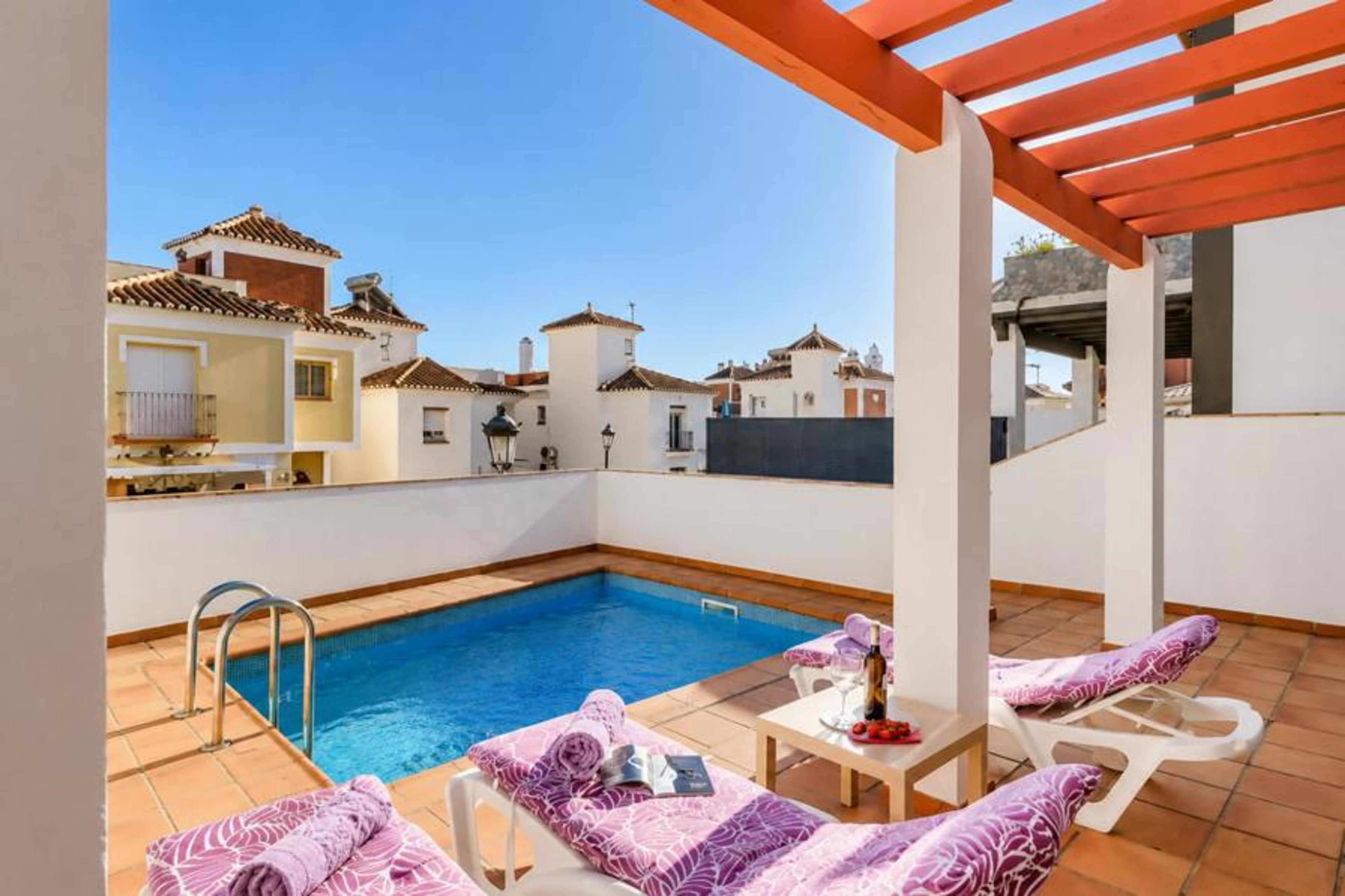 Private rooftop terrace at a modern vacation villa, showcasing a crystal-clear plunge pool, comfortable sun loungers with patterned purple cushions, a wooden pergola, and scenic views of a Mediterranean-style resort town.