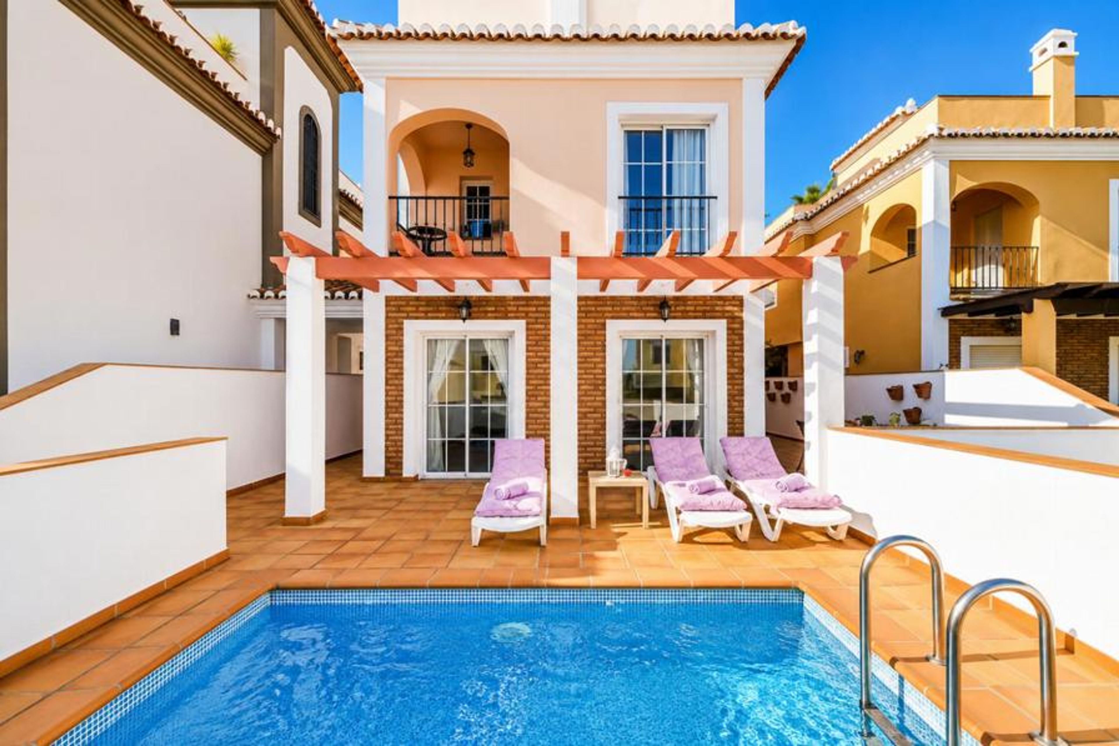 Luxury three-story self-catering townhome featuring a private plunge pool, a sun deck with purple sun loungers under a wooden pergola, and elegant arched balconies against a clear blue sky.